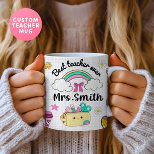 May include: White ceramic mug with the text "Best teacher ever" and "Mrs. Smith" in black script. The mug features a rainbow, clouds, and school-themed illustrations. A pink circle above the mug reads "CUSTOM TEACHER MUG".