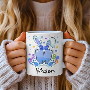 May include: White ceramic mug featuring a blue bunny design with the name "Mason" in black script. The design includes a blue bow tie, Easter eggs, and colorful stars. The mug is held by a person wearing a white sweater.