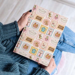 May include: A hardcover book featuring a repeating pattern of postage stamp-style illustrations. The design includes images of white geese, blue teapots, brown bread boxes labeled BREAD, pink lamps, and white casserole dishes on a light background.