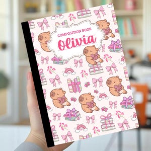 Puede incluir: Un cuaderno de composición con lomo negro y portada blanca que presenta un patrón repetitivo de capibaras de color marrón claro leyendo libros, lazos rosas, pilas de libros y pequeños arcoíris. El centro tiene una etiqueta con el texto Olivia.
