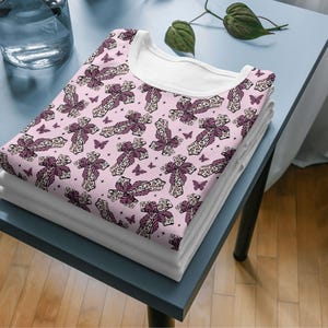 May include: A stack of folded white t-shirts with a pink patterned t-shirt on top. The pink shirt features a repeating pattern of crosses with bows and butterflies. The t-shirts are on a blue table.
