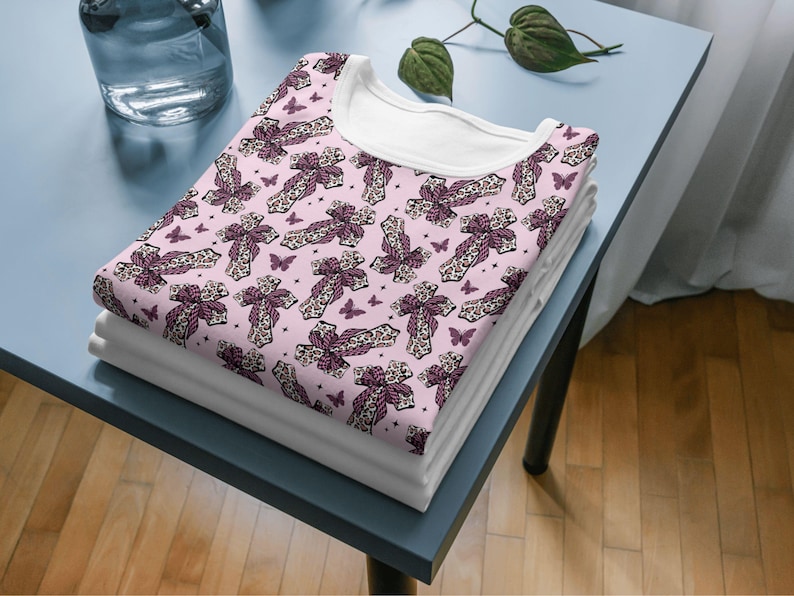 May include: A stack of folded white t-shirts with a pink patterned t-shirt on top. The pink shirt features a repeating pattern of crosses with bows and butterflies. The t-shirts are on a blue table.