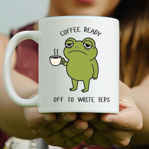 Könnte beinhalten: Weißer Keramikbecher mit blauem Innenraum, mit einer Cartoon-Frosch, der eine Kaffeetasse hält. Der Becher trägt den Text "COFFEE READY" über dem Frosch und "OFF TO WRITE IEPS" darunter.