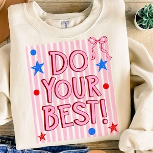 以下が含まれることがあります： ピンク色の「DO YOUR BEST！」の文字がプリントされたクリーム色のスウェットシャツ。ピンクと白のストライプの背景に、青い星、赤いドット、ピンクのリボンがデザインされています。カジュアルで快適なトップス。