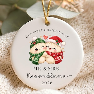 Puede incluir: Un adorno navideño redondo de cerámica blanca con un cordón dorado. El diseño muestra dos dumplings de dibujos animados con gorros y bufandas de invierno, con el texto "OUR FIRST CHRISTMAS AS MR.&MRS. Mason&Emma 2026" impreso en negro.
