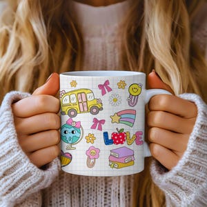 May include: White ceramic mug featuring colorful school-themed illustrations, including a yellow school bus, a globe, and the word "LOVE". The mug has a grid pattern background and is held by a person wearing a white sweater.