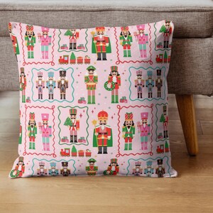 May include: A decorative pillow with a repeating pattern of colorful nutcracker figures in various uniforms, set against a light pink background. The design includes toy trains and Christmas trees, framed by a wavy border.
