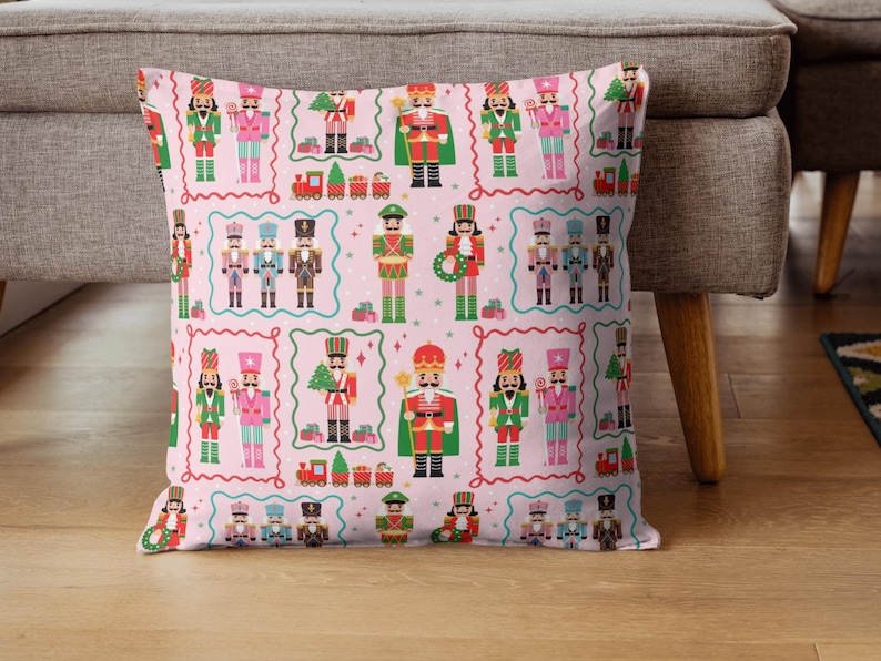 May include: A decorative pillow with a repeating pattern of colorful nutcracker figures in various uniforms, set against a light pink background. The design includes toy trains and Christmas trees, framed by a wavy border.