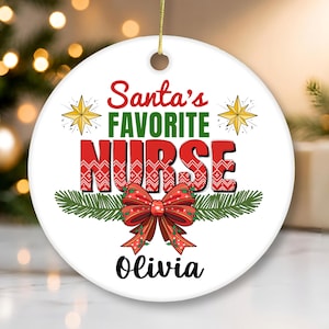 Puede incluir: Un adorno navideño redondo de cerámica blanca con el texto en rojo y verde Santa's FAVORITE NURSE en una fuente roja estampada, dos estrellas amarillas, ramas de pino verdes, un lazo rojo grande y el nombre Olivia impreso en negro abajo.