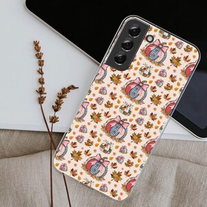 May include: A phone case with a fall-themed pattern. The design features pumpkins with bows, autumn leaves, and small stars on a cream background. The case is made of a clear material.