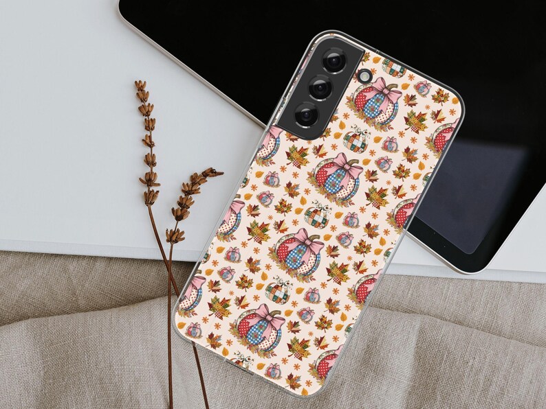 May include: A phone case with a fall-themed pattern. The design features pumpkins with bows, autumn leaves, and small stars on a cream background. The case is made of a clear material.