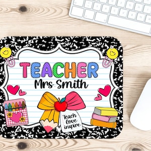 May include: A rectangular mousepad with a black and white speckled border. The center features a lined paper design with the word "TEACHER" in colorful letters and "Mrs. Smith" below. A pencil, apple, and bow graphic is present, along with hearts, crayons, and books.