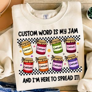 Puede incluir: Sudadera color crema con el texto "CUSTOM WORD IS MY JAM AND I'M HERE TO SPREAD IT!" con botes de mermelada coloridos con la palabra "Custom" en cada uno. El diseño incluye un patrón de cuadros blancos y negros.