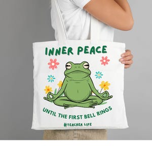 Puede incluir: Bolso tote blanco con una rana verde en postura de yoga, rodeada de flores. El texto "INNER PEACE" y "UNTIL THE FIRST BELL RINGS" están impresos encima y debajo de la rana, con "#TEACHER LIFE" en la parte inferior.