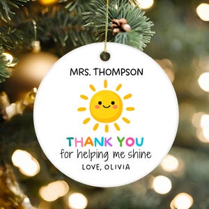Puede incluir: Un adorno redondo de cerámica blanca con un gráfico de sol amarillo con cara sonriente. El texto dice MRS. THOMPSON, THANK YOU for helping me shine y LOVE, OLIVIA. El adorno cuelga de un cordón dorado sobre un fondo desenfocado.