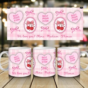 Puede incluir: Tazas de cerámica rosas con un diseño de "Mejor Mamá del Mundo", corazones, cerezas y lazos. Las tazas tienen los nombres Olivia, Madison y Thomas. Se muestra una pancarta a juego.