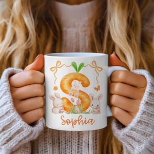 May include: White ceramic mug with an Easter design. The design features a large orange carrot-shaped letter "S" with a bunny inside, other bunnies, flowers, and the name "Sophia" in orange script. The mug is held by a person.