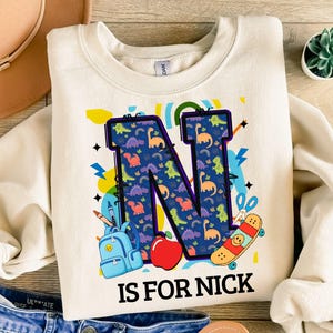 Puede incluir: Sudadera color crema con una gran letra "N" llena de un patrón de dinosaurios. El diseño incluye una mochila, una manzana, una patineta y el texto "IS FOR NICK".