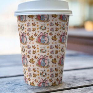 May include: A disposable coffee cup with a white lid. The cup is covered in a repeating pattern of pumpkins with bows, fall leaves, and stars in shades of pink, blue, red, and yellow on a cream background.