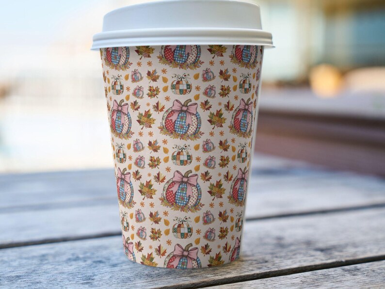 May include: A disposable coffee cup with a white lid. The cup is covered in a repeating pattern of pumpkins with bows, fall leaves, and stars in shades of pink, blue, red, and yellow on a cream background.