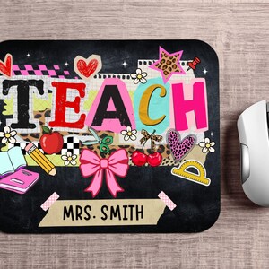 May include: Black rectangular mousepad with the word "TEACH" in colorful letters and school-themed graphics. Includes a pink bow, apple, pencil, and the name "MRS. SMITH". A white computer mouse is to the right.