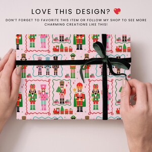 May include: A pink gift box with a repeating pattern of toy nutcracker soldiers, trains, and Christmas trees. The box is tied with a black ribbon and a dark green bow. The text "LOVE THIS DESIGN?" is at the top.