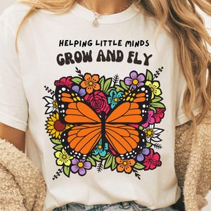 Puede incluir: Camiseta blanca con un diseño de mariposa colorida y el texto "HELPING LITTLE MINDS GROW AND FLY". La mariposa tiene alas naranjas y está rodeada de flores multicolores.