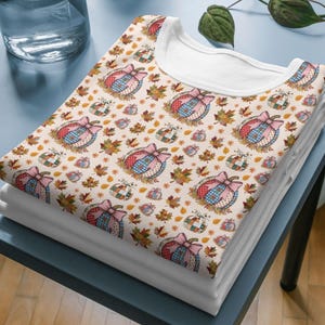 May include: A stack of white t-shirts with a repeating pattern of pumpkins, bows, and autumn leaves. The pumpkins have a patchwork design with pink, blue, and red accents. The leaves are in shades of brown and orange.