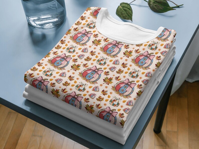 May include: A stack of white t-shirts with a repeating pattern of pumpkins, bows, and autumn leaves. The pumpkins have a patchwork design with pink, blue, and red accents. The leaves are in shades of brown and orange.