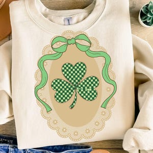 Puede incluir: Sudadera de color crema con un diseño de trébol a cuadros verdes y blancos dentro de un marco ovalado decorativo. El marco está adornado con un lazo y una cinta verdes, y un borde de encaje festoneado. Ropa temática del Día de San Patricio.