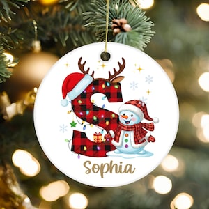 Puede incluir: Un adorno navideño redondo de cerámica blanca con una letra S grande en patrón de cuadros rojos y negros, decorado con un gorro de Papá Noel, astas de reno y luces de colores. Un muñeco de nieve sonriente lleva una bufanda a cuadros a juego.