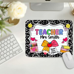 May include: A rectangular mousepad with a black and white composition notebook design. The mousepad features the text "TEACHER Mrs Smith" in colorful letters, along with illustrations of a bow, crayons, hearts, and books. A white computer mouse is next to the mousepad.