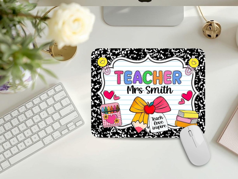 May include: A rectangular mousepad with a black and white composition notebook design. The mousepad features the text "TEACHER Mrs Smith" in colorful letters, along with illustrations of a bow, crayons, hearts, and books. A white computer mouse is next to the mousepad.