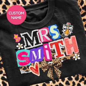 Puede incluir: Camiseta negra con un diseño gráfico colorido que dice "MRS SMITH". El diseño incluye un lápiz con un lazo, un corazón y un lazo con estampado de leopardo. Un círculo rosa en la parte superior dice "CUSTOM NAME".
