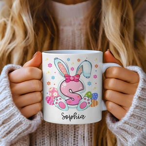 Puede incluir: Taza de cerámica blanca con un diseño de conejito rosa, un lazo rosa, la letra "S" y el nombre "Sophia". La taza tiene coloridos huevos de Pascua y flores. Un regalo personalizado para Pascua.