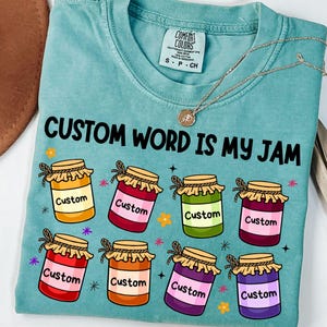 Puede incluir: Una camiseta turquesa Comfort Colors con el texto "CUSTOM WORD IS MY JAM" e ilustraciones de tarros de mermelada. Cada tarro tiene un color diferente y la palabra "Custom". Un collar de oro está sobre la camiseta.