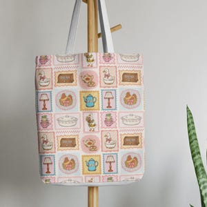 May include: A white tote bag featuring a repeating grid pattern of illustrations including geese, teapots, lamps, bread boxes, and baskets. The design uses a soft color palette of pink, blue, and yellow, with each icon framed in a scalloped border.