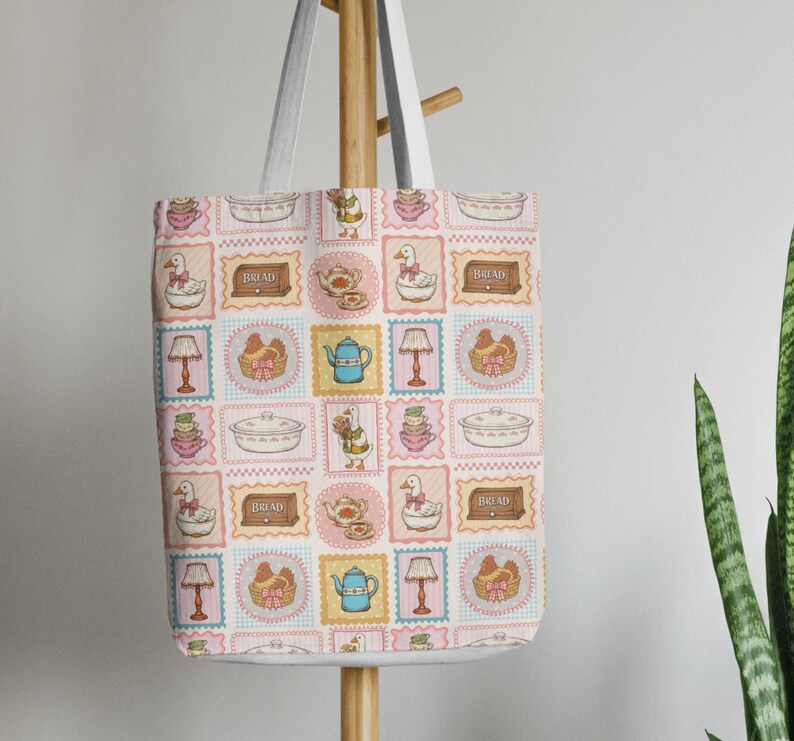 May include: A white tote bag featuring a repeating grid pattern of illustrations including geese, teapots, lamps, bread boxes, and baskets. The design uses a soft color palette of pink, blue, and yellow, with each icon framed in a scalloped border.