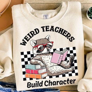 May include: Cream sweatshirt featuring a raccoon wearing red glasses, reading a book, and the text 'WEIRD TEACHERS Build Character'. The design includes a black and white checkered background. The sweatshirt is a neutral colour.