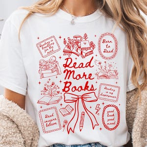 Puede incluir: Camiseta blanca con un diseño gráfico rojo que presenta el texto "Read More Books" e ilustraciones relacionadas con libros. El diseño incluye las frases "Reading is breathing for the mind", "Born to Read" y "Reading is my superpower".
