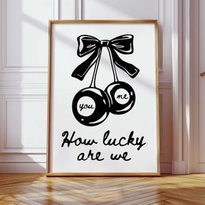 Puede incluir: Una impresión artística enmarcada con una ilustración en blanco y negro de dos cerezas atadas con un lazo. Las palabras "you" y "me" están en las cerezas, con la frase "How lucky are we" debajo. Decoración.