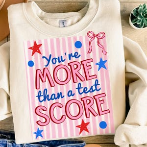 May include: Cream-colored sweatshirt with the text "You're MORE than a test SCORE" in pink and blue. The design includes pink stripes, stars, and a bow. The sweatshirt is folded on a wooden surface.