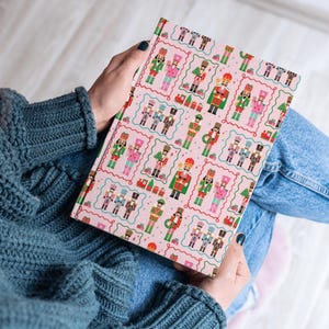 May include: A pink journal featuring a repeating pattern of colorful nutcracker soldiers in various uniforms. The journal is held open, displaying the patterned cover. The background includes a person wearing a blue sweater and jeans.