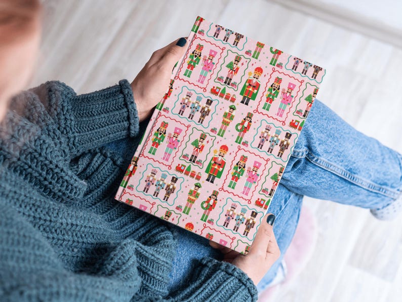 May include: A pink journal featuring a repeating pattern of colorful nutcracker soldiers in various uniforms. The journal is held open, displaying the patterned cover. The background includes a person wearing a blue sweater and jeans.