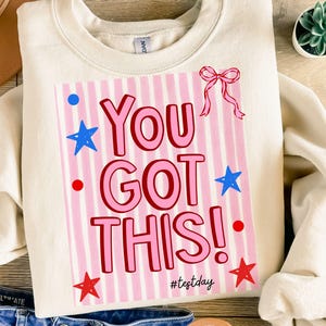 Puede incluir: Sudadera color crema con el texto "You Got This!" en rosa, sobre un fondo a rayas rosas y blancas. El diseño incluye estrellas azules, puntos rojos y un lazo rosa. El hashtag #testday está en la parte inferior.