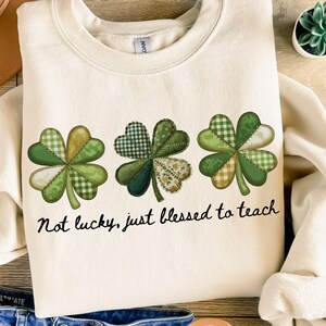 Puede incluir: Sudadera color crema con tres diseños de tréboles en tonos verdes con patrones de cuadros y florales. El texto "Not lucky, just blessed to teach" está impreso debajo. La sudadera tiene cuello redondo y mangas largas.