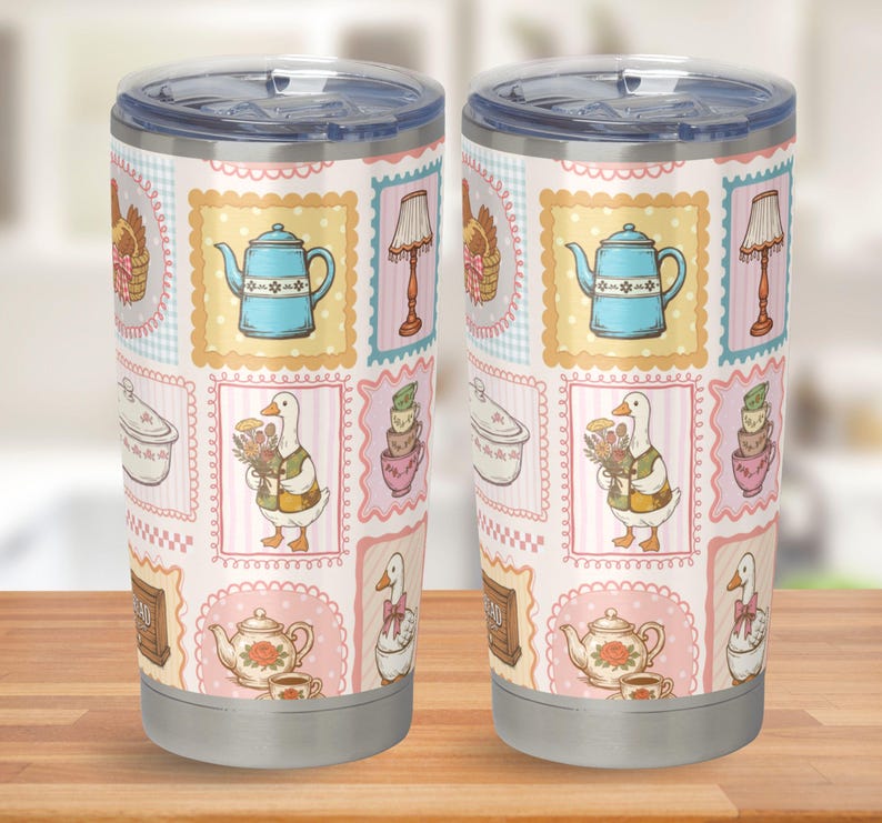 May include: Two stainless steel tumblers with clear plastic lids, featuring a repeating pattern of rectangular frames containing illustrations of blue teapots, geese, floral teapots, lamps, and stacks of teacups on a light pink background.