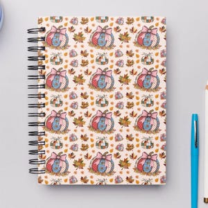 May include: Spiral-bound notebook with a fall-themed pattern. The design features pumpkins with pink bows, surrounded by autumn leaves on a cream background. A blue pen and a white pencil are next to the notebook.