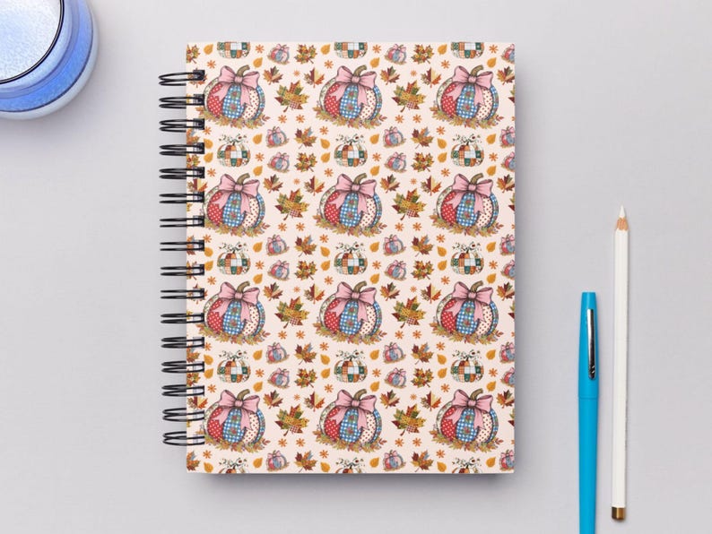 May include: Spiral-bound notebook with a fall-themed pattern. The design features pumpkins with pink bows, surrounded by autumn leaves on a cream background. A blue pen and a white pencil are next to the notebook.