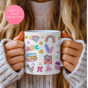May include: White ceramic mug decorated with colorful school-themed illustrations, including a rainbow, crayons, books, and the letters "ABC". The mug is held by a person wearing a beige sweater. A pink circle with the text "ADD TO YOUR FAVORITES!" is in the background.